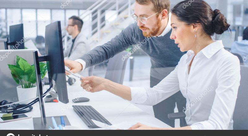 Beautiful Businesswoman Uses Desktop Computer, Consults Her Project Manager about Documents with Graphs and Statistics. In the Background Big Corporate Firm Office